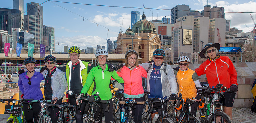 8 cyclists stand next to their bikes in front of the Flinders Railway Station dome. A photo of Sarah Maddock in 1894 has been placed on a video screen on one of the buildings in the background.