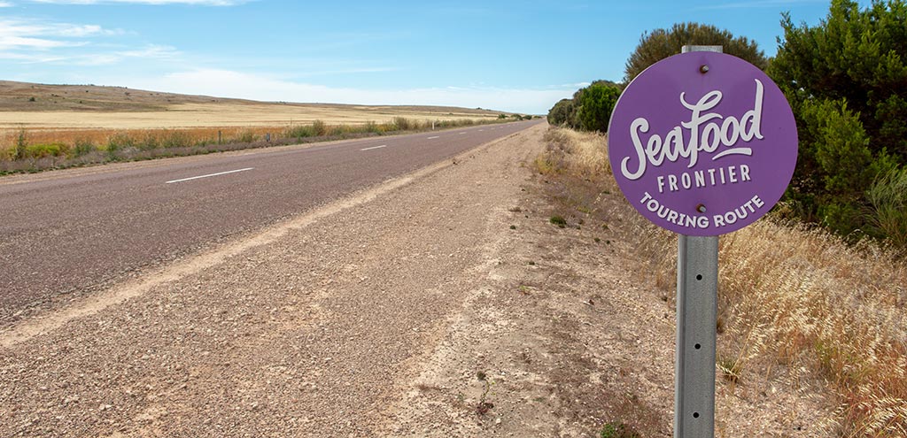 Purple disc shaped sign beside the Flinders Highway. The sign reads “Seafood Frontier Touring Route”.